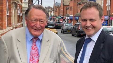 Sir John Stanley and Tom Tugendhat, both wearing formal suits, stand side by side in a street in Tonbridge, smiling into the camera.