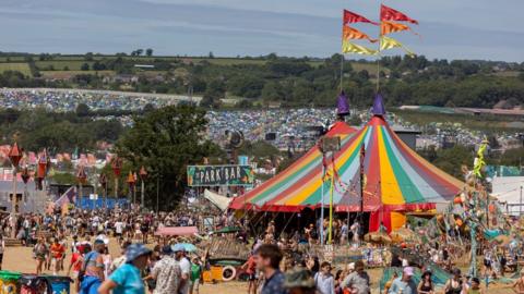A multi-coloured big top tent surrounded by people and a view across fields of tents at Glastonbury