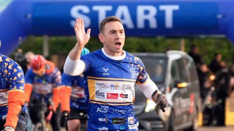 A man mid run, wearing a blue top, waves to a crowd. Cyclists behind him. A blue banner saying START is just behind him.