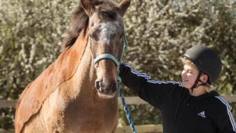 A boy is petting a brown horse. The boy is smiling and wearing a black jumpsuit and helmet.