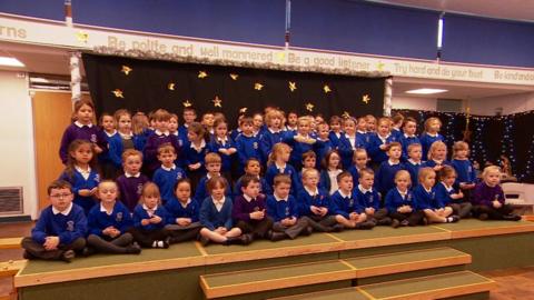 A stage in a school hall with steps leading up to the stage. One row of primary school ages children, some in blue jumpers and some in purple jumpers and in school uniform, sat on the stage. The row of children behind them are kneeling down on the stage. There are two rows of children stood up. The camera is angled so the children are looking to the right of the camera. Behind the stage is a black background with gold stars.