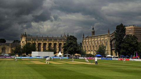 A wide view of cricketers from Gloucestershire and Hampshire playing during the Cheltenham Cricket Festival, with historic buildings in the background. The clouds are dark overhead