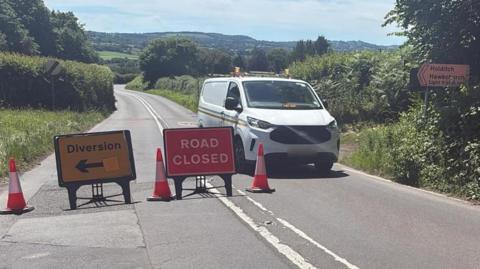 A white van parked on the road near the scene of a crash in the countryside. Road closed and diversion signs are on the road along with three traffic cones.