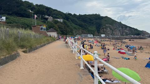 Greve de Lecq beach to the right with a path with a white railing on the left, many people are sitting, lying and walking on the sand under umbrellas and tents. To the left, grassy dunes and a small brown lifeguard hut with flags stand in front of tree-covered hills. 