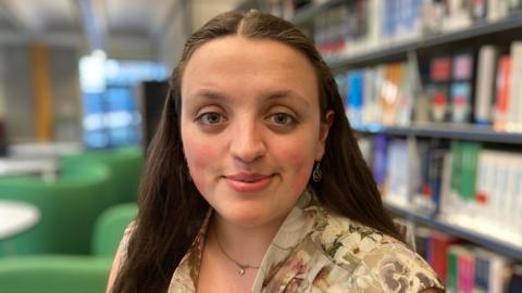 Verity Verhoven is looking straight into camera and in the background you can see a library area at the University of Derby. 