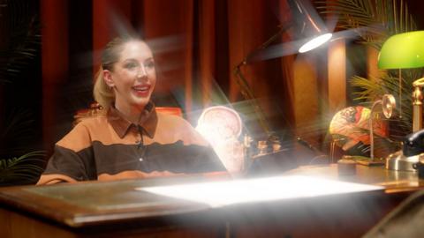 Katherine Ryan sits at a desk smiling while light sparkles around her