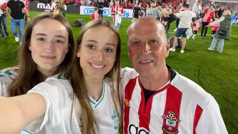 Kevin Screech on the right with his daughters. They are on the pitch surrounded by other fans at a Southampton FC game. They are all wearing Saints tops.