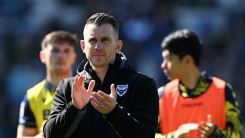 Matt Bloomfield applauding in a black Oxford United jacket with his players behind him