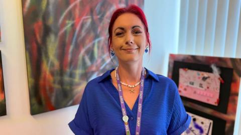 A woman with red hair, wearing a blue top. Ahe is looking at the camera smiling. She has a lanyard round her neck that says Staff on it. There are pictures and a closed vertical window blind behind her.