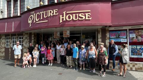 A large group of people outside a building called The Picture House. Many of them hold signs saying 'Save Our Cinema'.