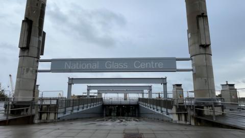 The entrance of the National Glass Centre in Sunderland. A large grey sign is suspended between two high cement pillars. A sloped path leads to the glass doors of the building. The fenced off roof of the building is visible in the distance.