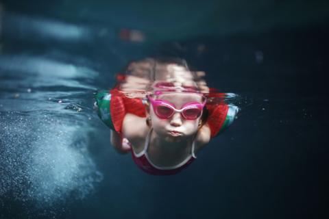photograph shows a girl swimming underwater wearing brightly coloured arm bands a swimsuit and goggles