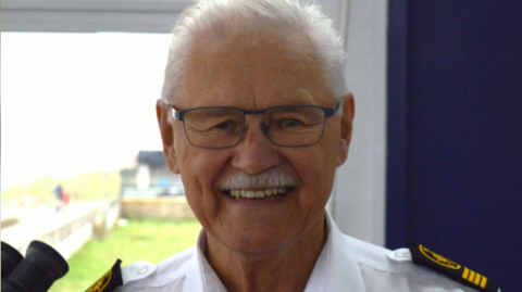 A headshot of a man with grey hair and a grey moustache wearing glasses and smiling into the camera. He is wearing Coastguard uniform that is white with black and yellow shoulder pads. 