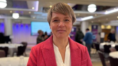 Alice Gill, a woman with short blonde hair and gold earrings. She is wearing a pink blazer over a white blouse. She is looking directly at the camera. Behind her is a conference room, with a large projector screen visible.