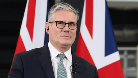 Sir Keir Starmer, dressed in a dark suit and light green tie, stands at a podium speaking. Behind him are two large Union Jack flags. His face is blurred in the image."