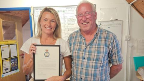 A picture of a man and a woman stood next to each other smiling at the camera. The woman on the left of the image is wearing a necklace, a white shirt and has blonde hair. She is holding a letter in a frame. The man on the right is wearing glasses, has grey hair and a blue pattern shirt.