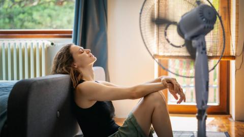 
Young woman sitting at home on the floor in front of a fan saving from heat - stock photo