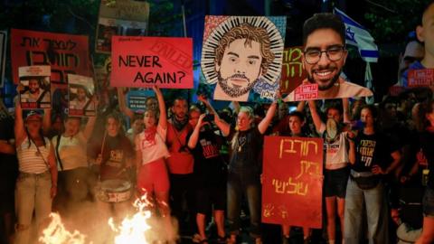 A crowd of protesters demanding the release of hostages hold signs in front of a fire in Tel Aviv