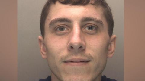 A police mugshot of a man in his early 20s, who has brown hair and light stubble. He is smiling at the camera. 