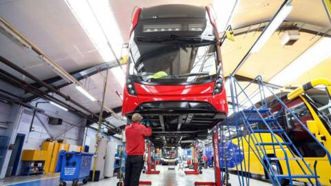 Worker dressed in black trousers and red top stands beneath a red bus at Alexander Dennis' factory in Eastfield.