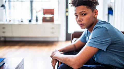 An anxious-looking teenager sits in a room, looking at the camera. 