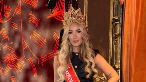 The image shows a blonde woman standing in a theatre box, wearing a crown and a bright red sash that reads "Ms Glamour UK 2025".