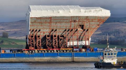 The front section of a large ship, carried by a blue barge with a tug in the foreground