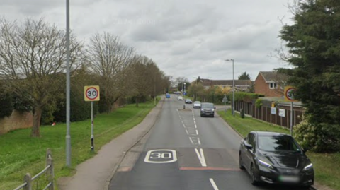 A Google Streetview image of a single-carriageway residential road. There is a 30mph speed limit sign. It is cloudy.