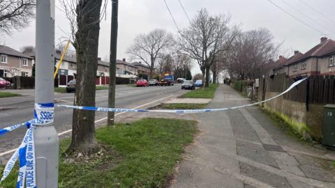 Police tape reading “POLICE LINE DO NOT CROSS” stretches across a suburban street, blocking access. Houses, parked cars, and bare trees line the road in the background on an overcast day.