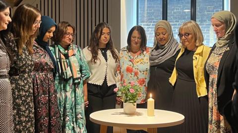 Muslim and six Jewish women standing around a table, looking at a lit candle and a potted miniature rose