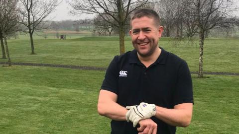 A man wearing a black polo-shirt while smiling at the camera. He is wearing white gloves while holding onto a golf club. He is standing in a field