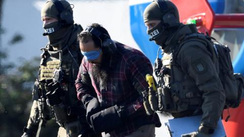 A bearded man, blindfolded, with ear protectors and his hands bound in heavy mitts, is escorted by heavily armed police in full tactial gear, faces hidden by balaclavas