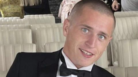 Ryan Willcox wears a dark suit and bow tie with a white shirt. He smiles at the camera and his head is shaved. Rows of white seats can be seen behind him.