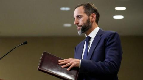 Jack Smith stands in front of a microphone, opening a leather-bound folder or notebook. He has dark hair and a beard.