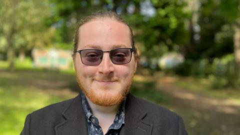 Steven Lewkowicz, 25, wearing tinted glasses, a beard, and hair in a pony tail, smiles at the camera in a park