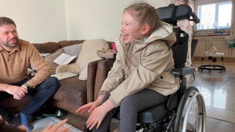 Smiling young girl in a wheelchair with her father looking on