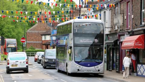 Bus drives through a York street with shoppers and shop awnings to the right, bunting above and other cars, vans and buses driving both ways down the street. 