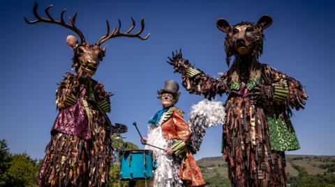 A giant stag costume, a giant bear costume and a woman in the middle dressed as a squirrel with a top hat banging a blue drum.