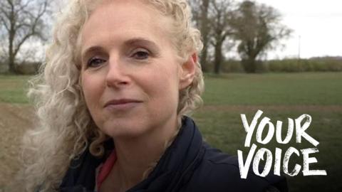 A woman with long, blonde, curly hair smiles at the camera. She is wearing a dark top with a pink collar. Behind her is a green field.