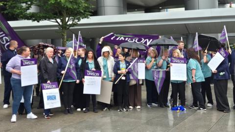 A group of hospital workers, holding signs and banners, outside a large hospital building 