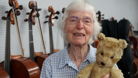 Jaquelin du Pre's sister smiles at the camera and holds an old teddy bear in front of a row of cellos