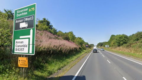 The A75 at Annan. A large sign on the left side of the image points to Stranraer and Dumfries ahead. It points to Annan and Canonbie to the left. 