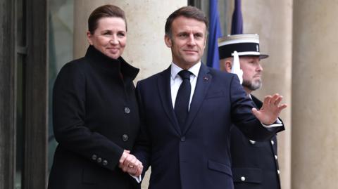 Mette Frederiksen, wearing a dark coat, shakes President Macron's hand. He is wearing a dark blue suit jacket and a tie.