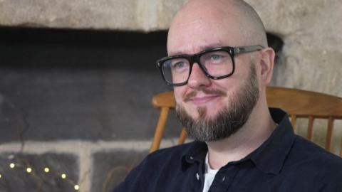 A man with a bald head and a brown beard and glasses, wearing a navy shirt over a white t shirt. He is sitting in a wooden chair and smiling. 