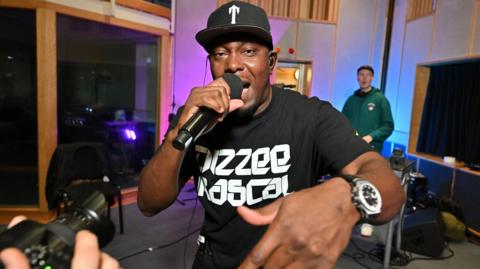 Dizzee Rascal looks at the camera and is wearing a t-shirt with the words Dizzee Rascal on it. He is holding a microphone in one hand and wearing a black baseball cap.