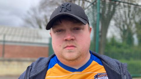 A man in a football shirt and cap looks at the camera. The shirt is orange and blue. Behind, green space is blurred.