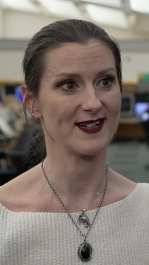 Amanda looks slightly off camera while giving an interview. She has light brown hair tied back in a ponytail. She has two necklaces with large dilver pendants and wears a cream knit jumper.