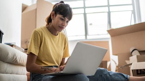 A person in a yellow top sits on their laptop at home with moving boxes behind them