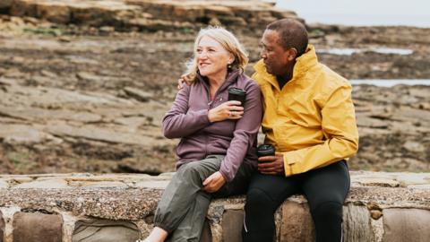 A middle-aged couple sit on a stone wall by a seaside. Both are holding coffee cups and are wearing bright coats.