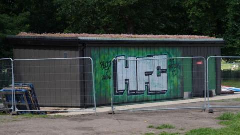 One wall of the toilet block has been spray painted green and there are three large letters on the side "HFC". There is a workmen's temporary fence around the block.